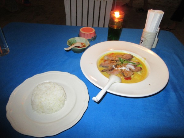 a table covered by a blue tablecloth with a plate of rice and a plate of excellent red duck curry, some condiments and a candle
