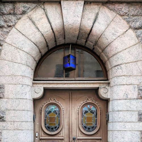 Painted brown door with 2 round stained glass windows set in a sturdy stone frame. There is a simple plain glass fanlight which contains a triangular blue panel that reads 15B in an Art Nouveau-style font.
