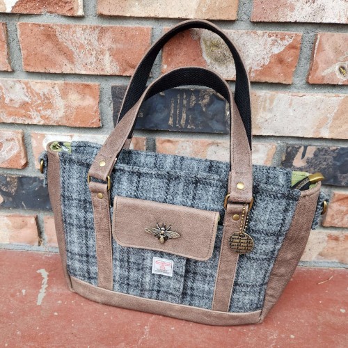 A purse leaning against a brown brick wall. The purse is a grey and black plaid Harris Tweed, it has a leather bottom, sides and straps. There is a leather flap pocket in the center of the front with a bee clasp. And there is a metal tag hanging from the handle hardware that has handmade stamped over and over in a delicate scroll.