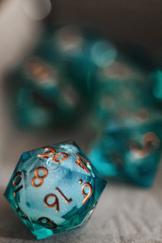 A turquoise translucent twenty sided dice sits in a dice tray, the numbers are inked in copper. It has a shallow depth of field showing other dice behind it that are out of focus. The dice has the number twenty facing up.