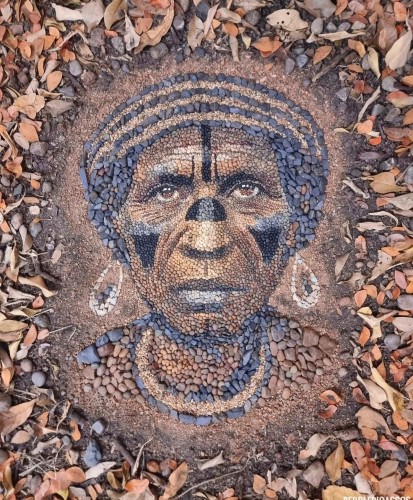 Urbanart. An impressive mosaic of small gray, brown, black and yellow pebbles surrounded by brown leaves. It is the portrait of an elderly, black, indigenous woman from the Amazon region. The elderly lady wears braided hair in parallel black and brown stripes. Her face is decorated with the typical black face painting. Triangles on the cheeks, a thin line across the forehead and a broad line across the nose. She wears square earrings and a wide, multi-colored necklace. The whole beautiful picture is made of pebbles. Title: "Jurema"
Info: "Jurema" is a female name sometimes attributed to members of the Guajajara tribe, also known as the "Guardians of the Amazon". The tribe is known for protecting the jungle from illegal deforestation.