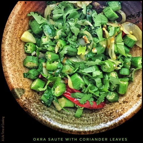 Sauteed sliced okra sit in a plate topped with coriander leaves. A red chilli can be seen amongst the okra, as well as onion, ginger and garlic.